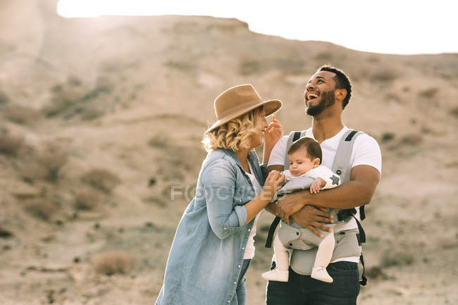 Feliz casual caucásico esposa y negro sonriente marido llevar bebé en la naturaleza en el día - foto de stock