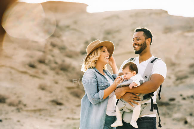 Feliz casual caucásico esposa y negro sonriente marido llevar bebé en la naturaleza en el día - foto de stock