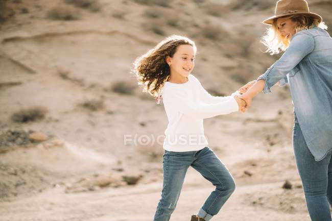Mujer rubia alegre girando con una hija casual sonriente y tomándose de la mano en el paisaje del desierto - foto de stock