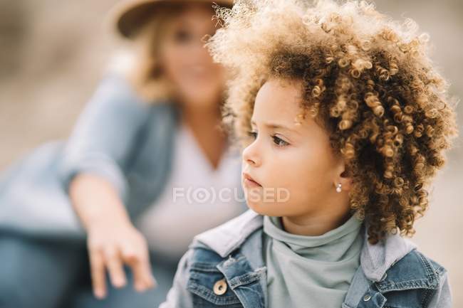 Concentrated serious toddler with curly hair sitting on nature and looking away while woman resting behind and smiling — Stock Photo