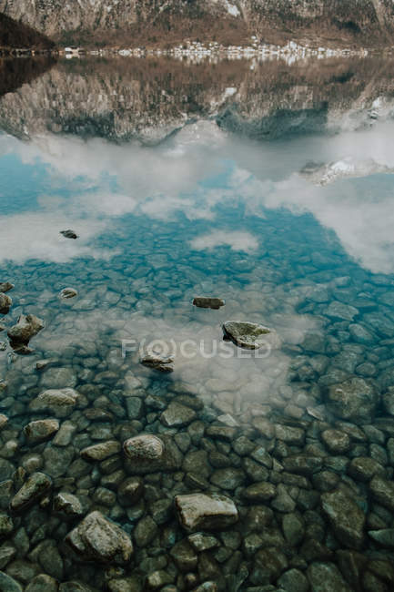 Von oben Steinufer und kristallklares Wasser, das den wolkenverhangenen Himmel in den Bergen in Hallstatt reflektiert — Stockfoto