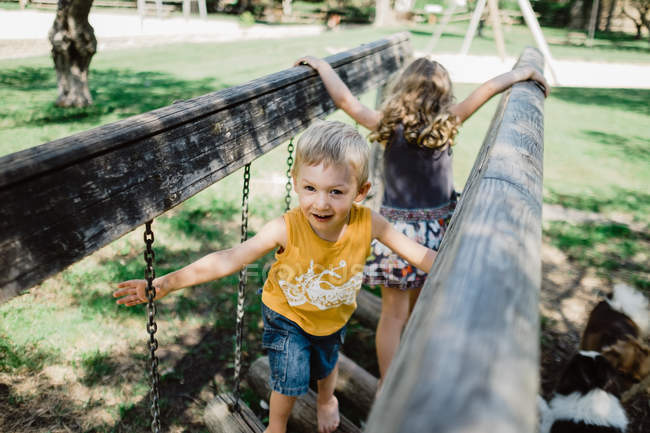 Fröhlicher Junge und begeistertes Mädchen klettern bei sonnigem Wetter spielend und auf Kettenhölzern auf Spielplatz auf grünem Rasen — Stockfoto