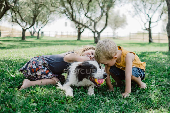 Liebliche Jungen und Mädchen umarmen flauschigen Hund mit Ball im Mund auf Gras — Stockfoto