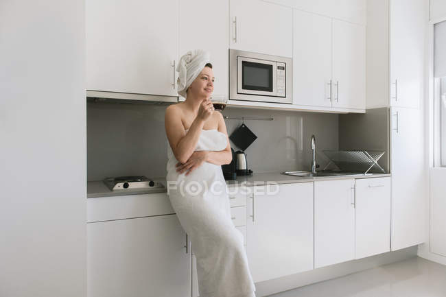 Mujer en toallas de baño blancas sonriendo y soñando mientras bebe café de pie en la cocina confortable contemporánea - foto de stock