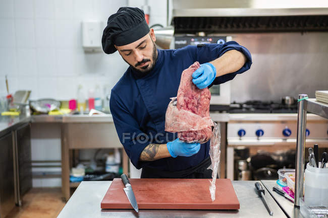 Male chef in dark blue uniform and gloves pulling large piece of raw meat out of vacuum plastic package — Stock Photo