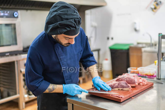 Cuoco maschio in uniforme e guanti in piedi a tavola in cucina professionale e affettatura di manzo sottilmente su tagliere in legno — Foto stock