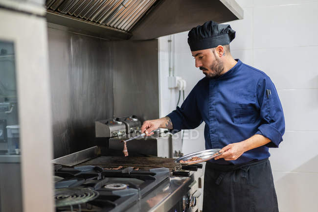 Cuoco maschio in grembiule nero e cappello in mano piatto di metallo e pinze e grigliate fette di carne sui fornelli mentre lavora in cucina ristorante — Foto stock