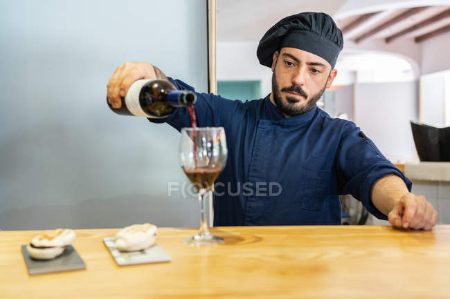 Cocinero masculino serio en uniforme azul y sombrero negro vertiendo vino tinto en vidrio mientras está de pie en el mostrador con sándwiches - foto de stock