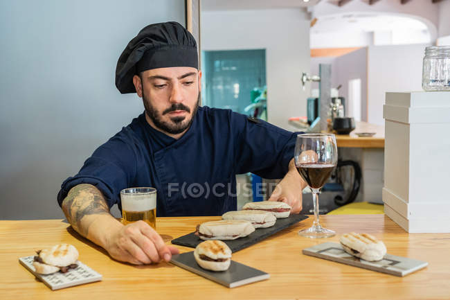 Серйозний професійний чоловік готує в уніформі і капелюсі аранжування лотка з гастрономічними сендвічами, стоячи за стійкою — стокове фото