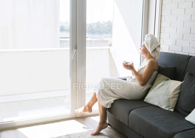 Woman in white towels looking out window wall and enjoying view while having refreshment with tasty beverage and sitting on soft couch — Stock Photo