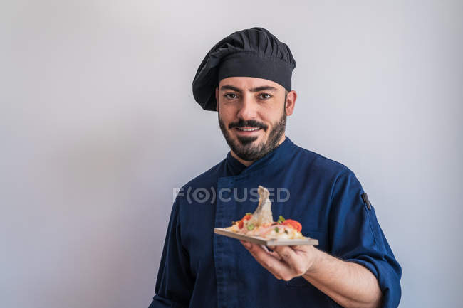 Щасливий дорослий бородатий шеф-кухар в темно-синій формі і чорний капелюх тримає в ручній тарілці з гастрономічною стравою — стокове фото