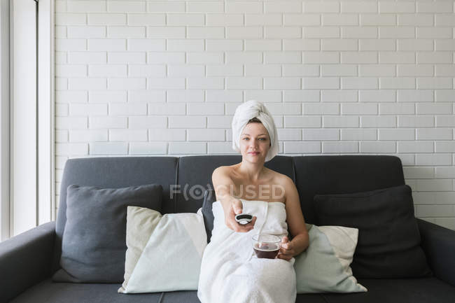 Happy relaxed woman enjoying day off while having hot drink after shower and watching TV in apartment in loft style — Stock Photo