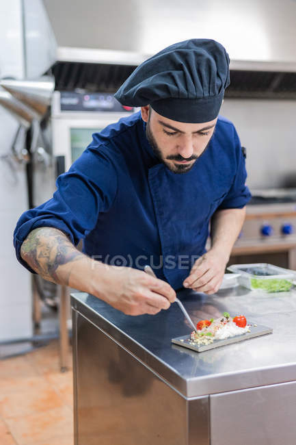 Cocinero masculino profesional concentrado que adorna y termina el delicioso plato listo para servir mientras trabaja en el mostrador de metal en la cocina del restaurante - foto de stock