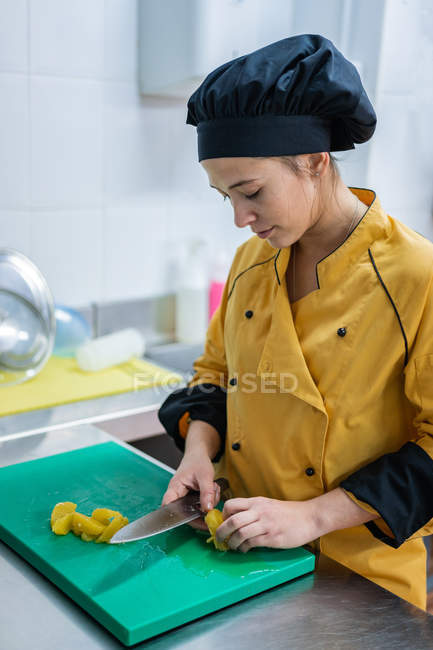 Giovane donna in uniforme gialla e cappello nero taglio succosa frutta gialla sul tagliere verde mentre lavora in cucina ristorante — Foto stock