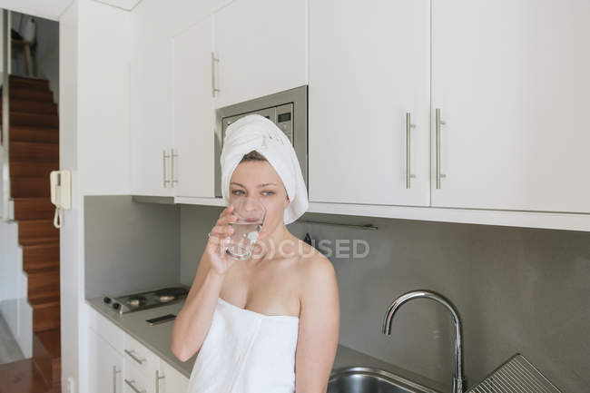 Жінка в білих рушниках для ванни дивиться в сторону і розмірковує, стоячи і спираючись на кухонні меблі і пиття — стокове фото