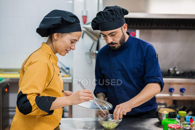 Jovem cozinheira feminina em amarelo uniforme segurando placa e pinças e colega masculino em azul escuro tigela uniforme segurando com alimentos — Fotografia de Stock