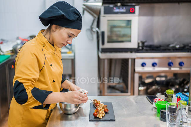 Side view of serious young female chef garnishing and finishing gourmet dish ready for serving while working at table — Stock Photo