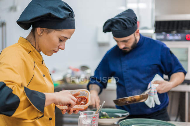 Vue latérale de la jeune cuisinière en uniforme jaune tenant pot avec sauce tout en travaillant avec un collègue masculin dans la cuisine du restaurant — Photo de stock