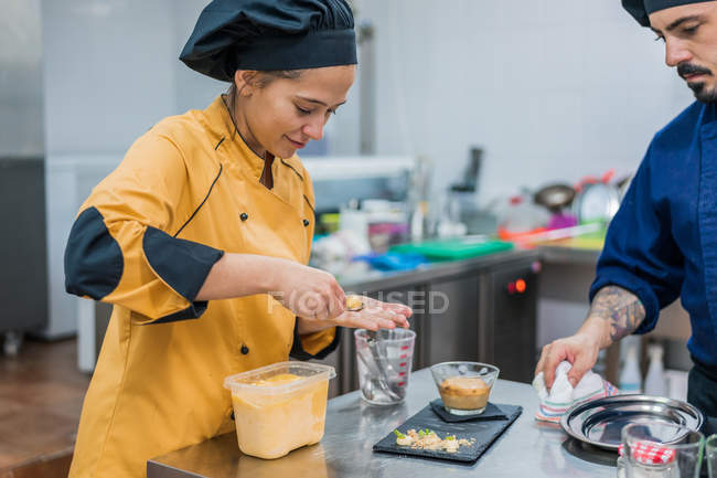 Чоловічий шеф-кухар спостерігає за молодою жінкою-асистенткою з ложкою в руці, кладе їжу на тарілку, працюючи разом на кухні ресторану — стокове фото