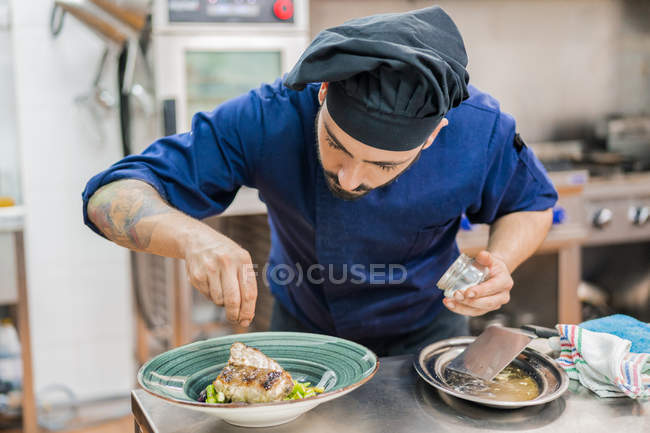 Cozinheiro macho em uniforme azul e chapéu preto prato de acabamento e colocando sal na placa de porção com peixe antes de servir — Fotografia de Stock