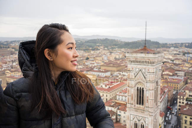 Mujer asiática feliz sonriendo y mirando hacia otro lado mientras está de pie en la cúpula de Brunelleschi contra las viejas calles de Florencia durante el viaje en Italia - foto de stock