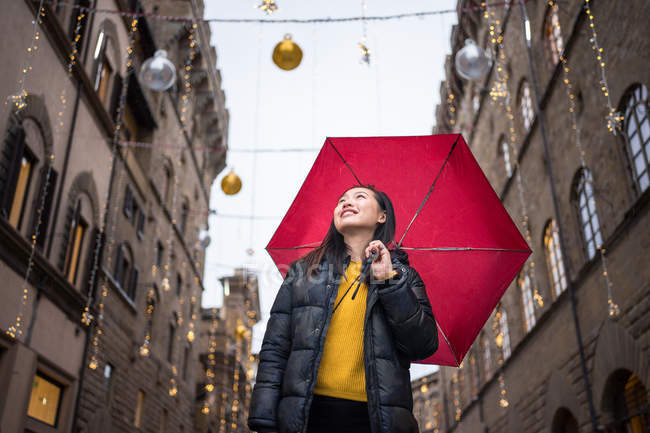 Basso angolo di donna felice con ombrello rosso sorridente e guardando in alto mentre camminava su una strada decorata invecchiata a Firenze, Italia — Foto stock
