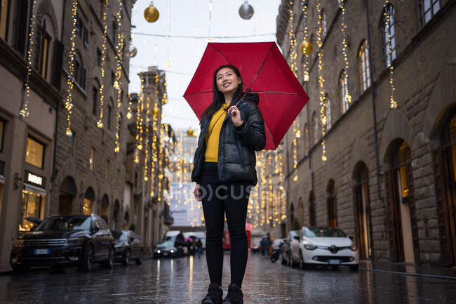 Bajo ángulo de mujer feliz con paraguas rojo sonriendo y mirando hacia arriba mientras camina en la calle decorada envejecida en Florencia, Italia - foto de stock