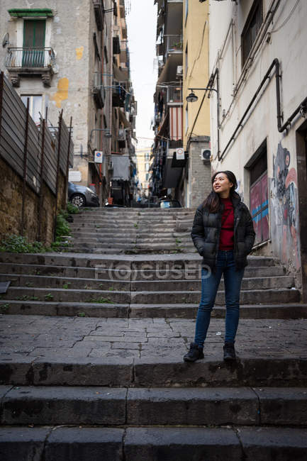 Asian woman with hands in pockets looking away while standing on stairs outside weathered buildings on city street — Stock Photo