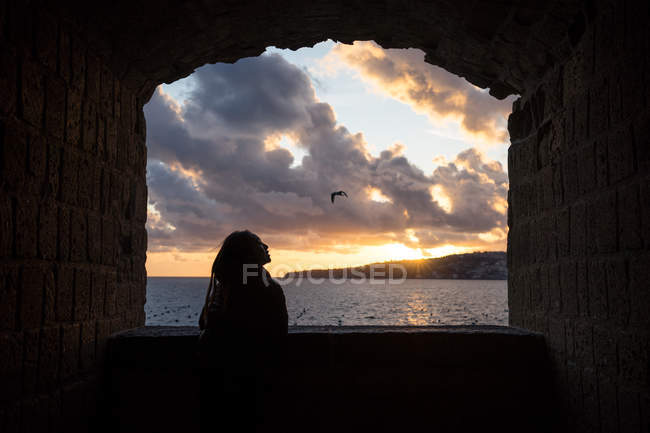 Обратный вид женщины в теплой одежде, наслаждающейся видом на красивый закат с облачным небом на берегу моря в Неаполе в Италии — стоковое фото