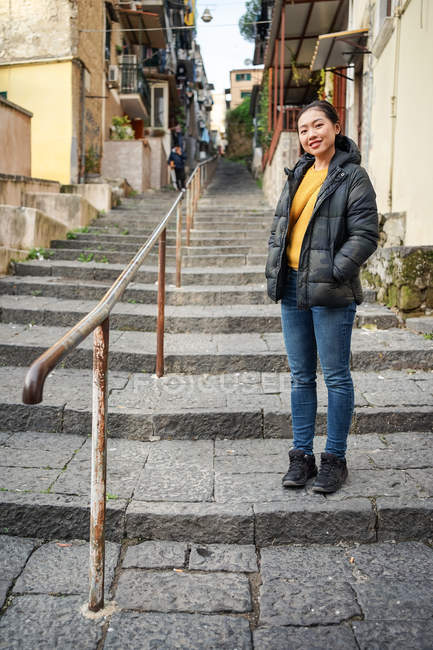 Mujer con las manos en los bolsillos mirando a la cámara mientras está de pie en las escaleras fuera de los edificios envejecidos en la calle de la ciudad - foto de stock