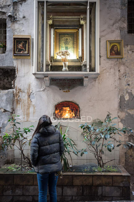 Vue arrière de la femme regardant le mur et la fenêtre altérés avec des icônes tout en se tenant sur la rue à l'extérieur de la vieille église — Photo de stock
