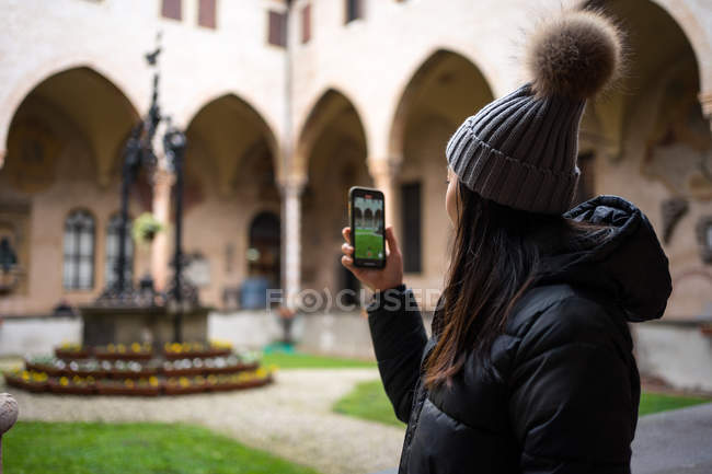 Вид сбоку на азиатскую туристку в теплой одежде и шляпе, фотографирующуюся на мобильный телефон во время посещения древнего города Сан-Антонио в Падуе в Италии — стоковое фото