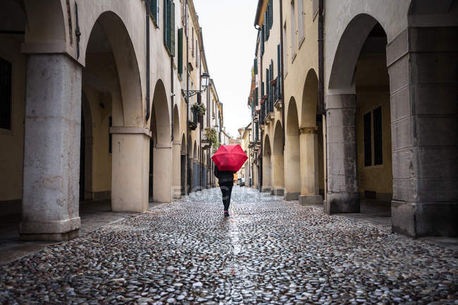 Vue arrière d'un voyageur méconnaissable en vêtements chauds en utilisant un parapluie rouge avec de vieux bâtiments sur fond flou à Padova en Italie — Photo de stock