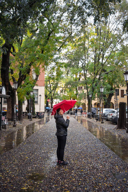 Joven viajera asiática en ropa de abrigo haciendo turismo usando paraguas rojo con edificios antiguos sobre fondo borroso en Padova en Italia - foto de stock