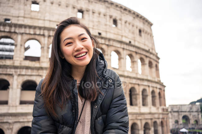 Desde abajo mujer feliz sonriendo y mirando a la cámara mientras está de pie sobre el fondo borroso del Coliseo en la calle de Roma, Italia - foto de stock
