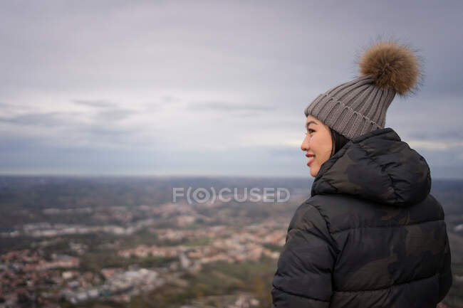 Vista posteriore della donna asiatica sorridente in cappello a maglia con pompon e giacca calda godendo della città in un posto incredibile a San Marino, Italia — Foto stock