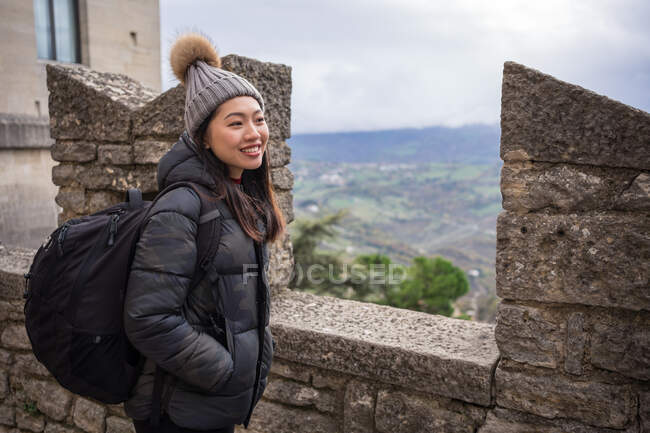 Вид сбоку улыбающейся азиатской женщины в вязаной шляпе с капюшоном, теплой куртке и рюкзаке, наслаждающейся городом в удивительном месте в Сан-Марино, Италия — стоковое фото