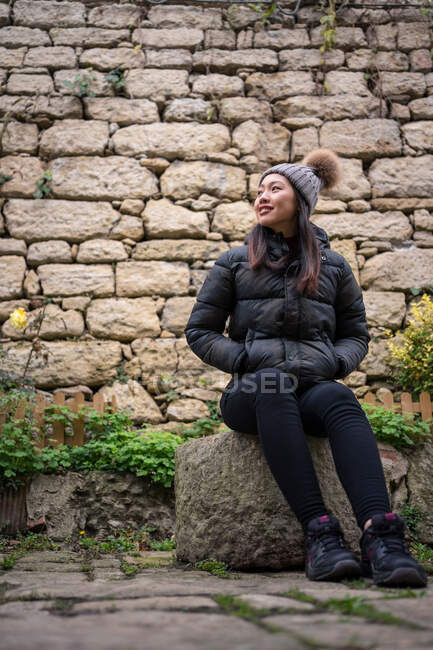 Mujer asiática alegre en sombrero de punto con pompón y chaqueta disfrutando sentado en piedra grande en el fondo de la pared de piedra en San Marino, Italia - foto de stock