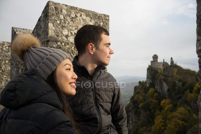 Nachdenklicher Mann und lächelnde Asiatin mit Strickmütze und Bommel, die wegschauen und die atemberaubende Landschaft des antiken Schlosses in San Marino, Italien, genießen — Stockfoto