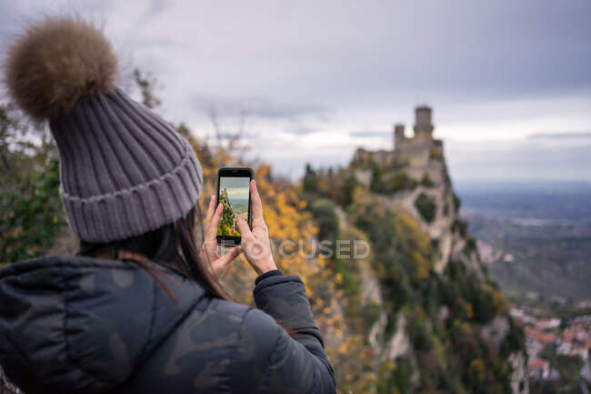 Обратный вид женщины в шляпе с помпоном и курткой, фотографирующей на мобильный телефон удивительного места в Сан-Марино, Италия — стоковое фото