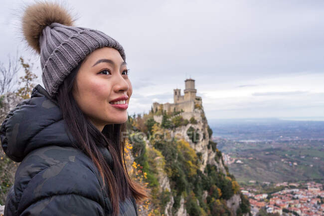 Vista lateral da mulher asiática em chapéu de malha com pompom e casaco desfrutando da cidade e da natureza em um lugar incrível em San Marino, Itália — Fotografia de Stock