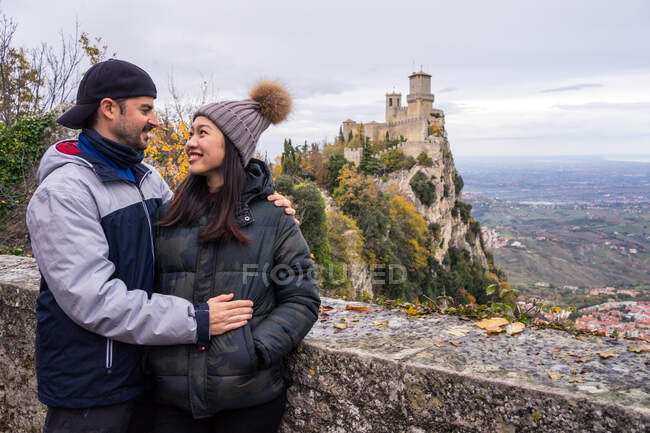 Homem amoroso abraçando sorridente mulher asiática em chapéu de malha com pompom inclinado sobre cerca de pedra na colina com paisagem incrível do castelo antigo em San Marino, Itália — Fotografia de Stock