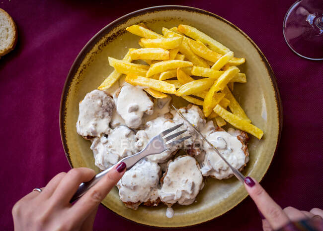 Vista superior de la señora de la cosecha con tenedor y cuchillo comiendo papas fritas con carne en salsa en el restaurante de Málaga en España - foto de stock
