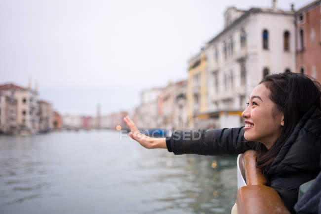 Vista lateral de la mujer asiática en reposo con ropa de abrigo sonriendo y extendiéndose desde el barco en la vía navegable en medio de edificios antiguos - foto de stock