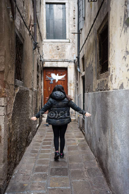 Back view of black haired faceless female in warm clothing walking with raised arms on narrow rocked old alley — Stock Photo