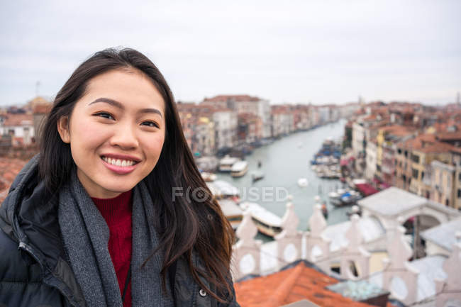Радісна азіатська жінка - мандрівниця в теплому одязі посміхається і дивиться на камеру з стародавнім містом і водними шляхами на невиразному тлі в хмарну погоду. — стокове фото