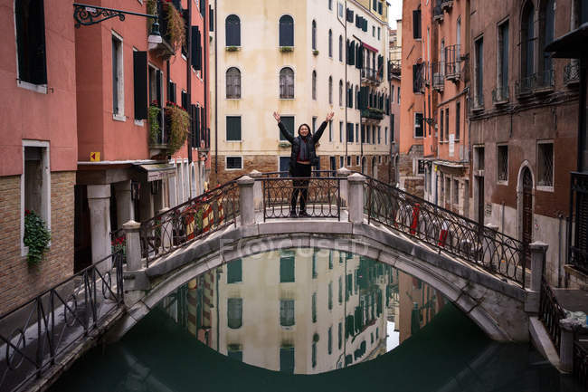 Content woman on vacation in warm clothing standing and raising hands on small bridge above water canal between old colorful buildings — Stock Photo