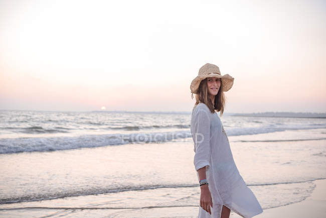 Розслаблена витончена жінка з довгим волоссям в капелюсі і світло-білою сорочкою на узбережжі під хвилястою водою на пляжі — стокове фото