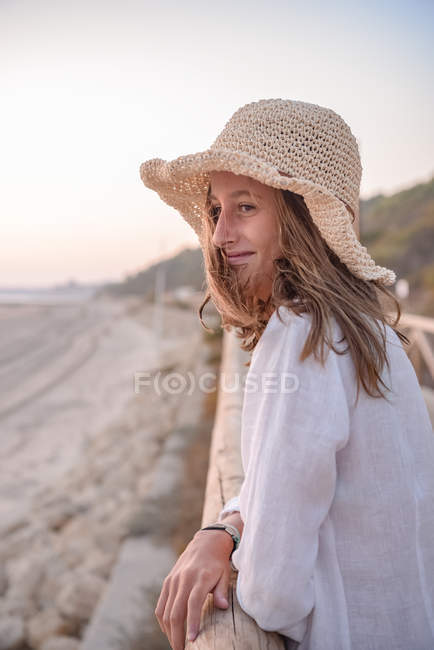 Encantadora mujer con sombrero apoyada en la cerca y mirando al mar - foto de stock