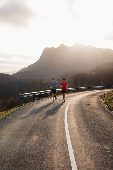 Active healthy male joggers running together on curved asphalt road with sunlight from behind mountain in background — Stock Photo